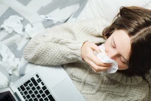 A young woman lies in bed with a laptop and blows her nose in a napkin. Seasonal cold or flu concept