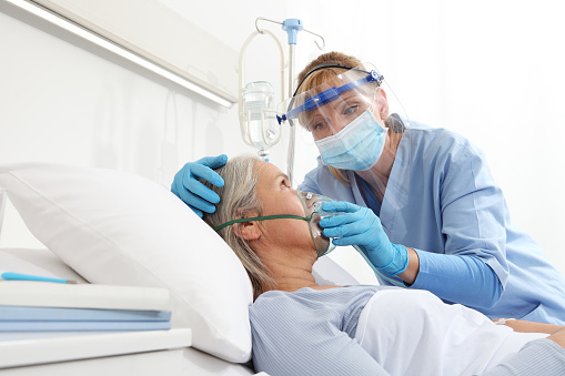 nurse puts oxygen mask on elderly woman patient lying in the hospital room bed, wearing protective gloves and visor medical mask, coronavirus covid 19 protection concept