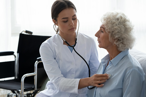 Young female physician caring about elderly disabled woman sit on couch using stethoscope listen sounds by heart or lungs, health test of older generation, medical insurance nursing eldercare concept
