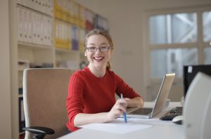 Woman holding a pen and sitting in front of a laptop while smiling brightly
