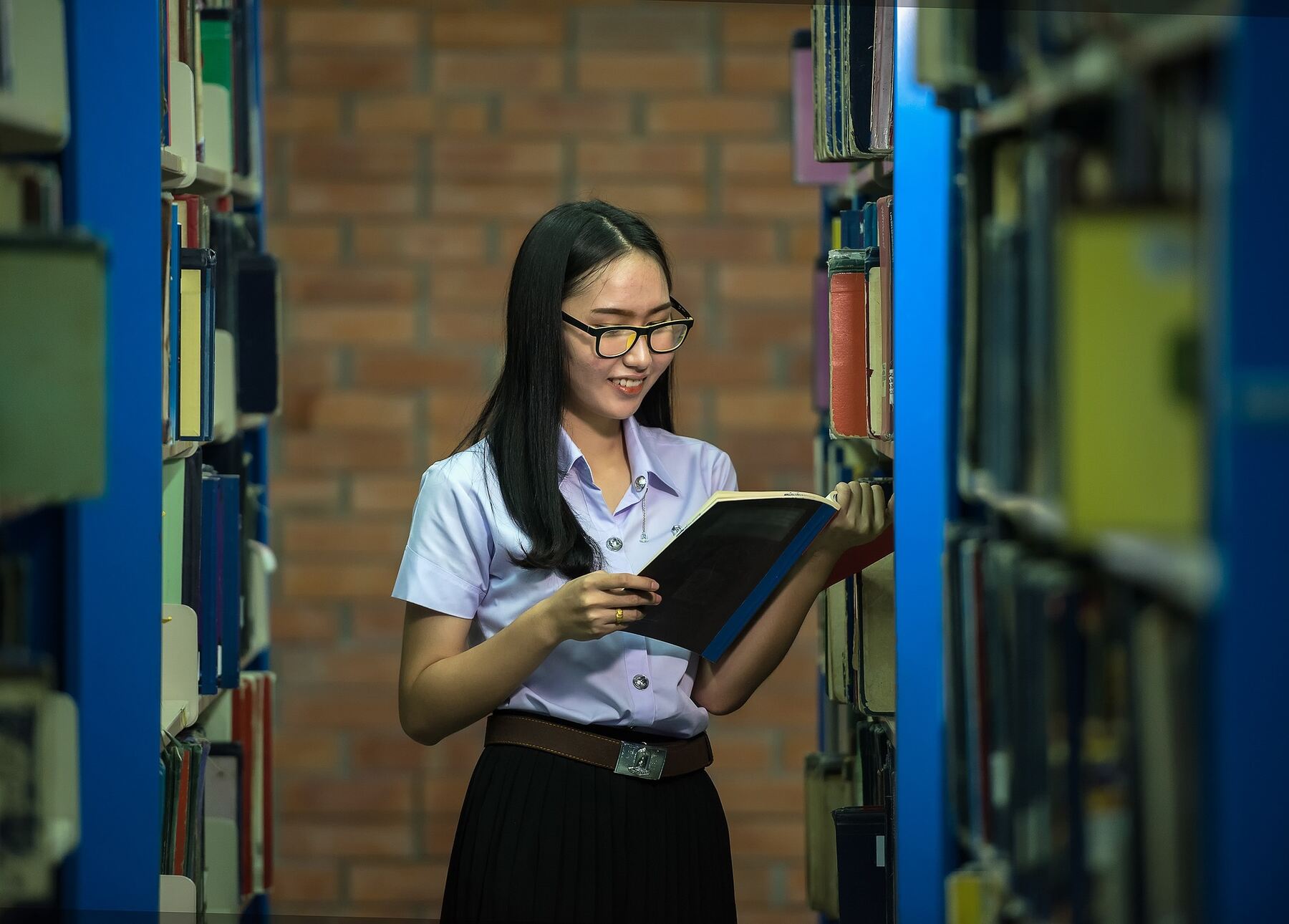 A young woman in glasses reading a book while standing in a library, surrounded by shelves of books