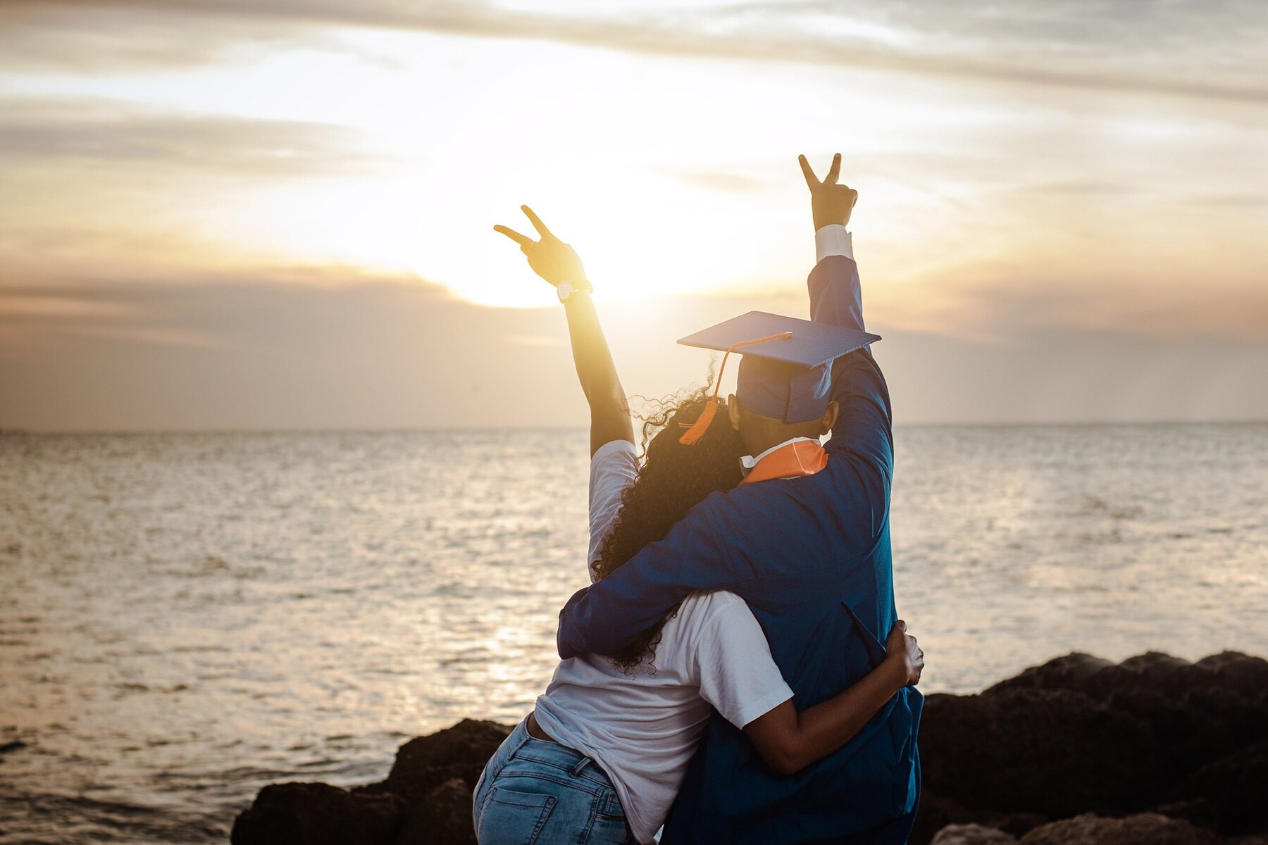 A woman and a man in graduation robes and cap celebrating on a beach with their arms around each other