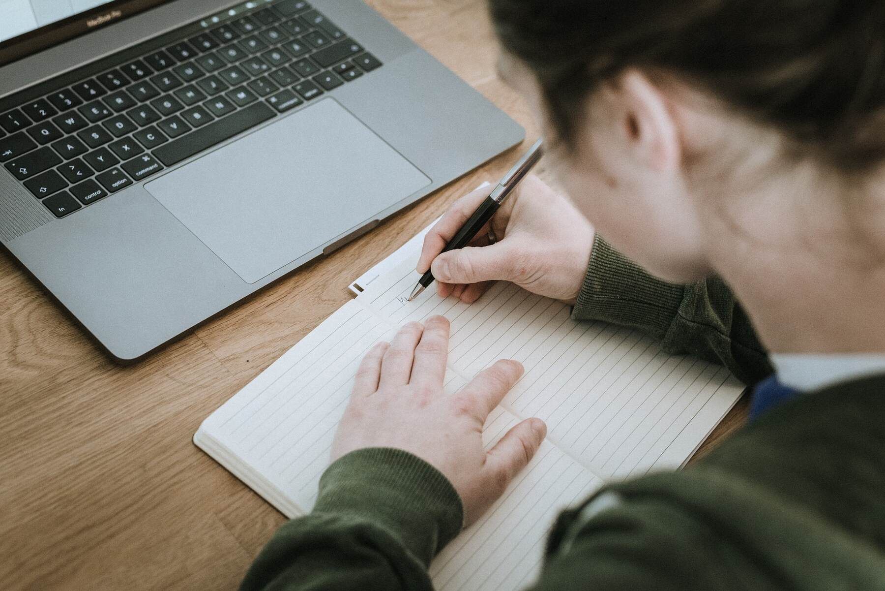 Woman jotting notes in notebook on wooden table with laptop nearby