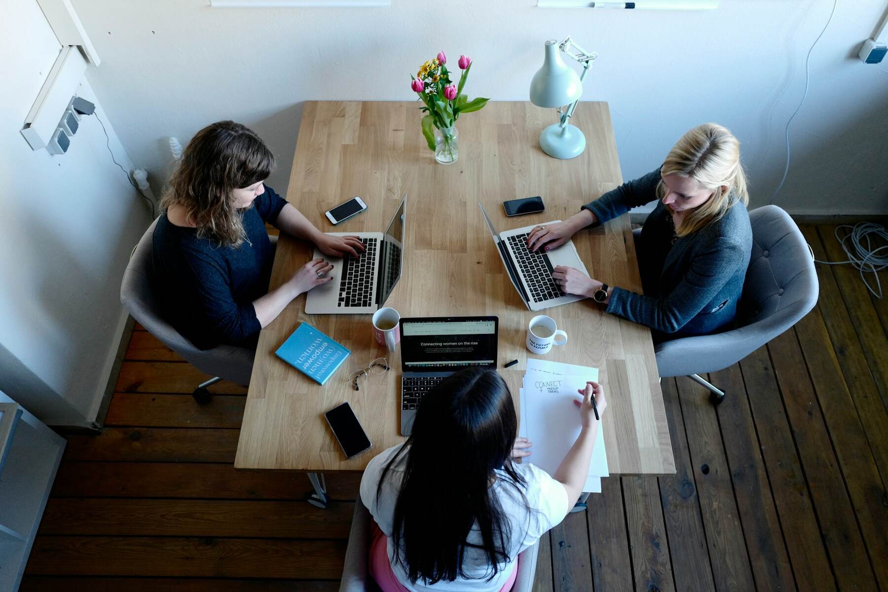 Three professional women sitting at a table with laptops, in a contemporary workspace